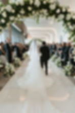 Bride and man walk down a flower-decorated aisle at an indoor wedding, surrounded by guests in formal attire, under soft lighting.