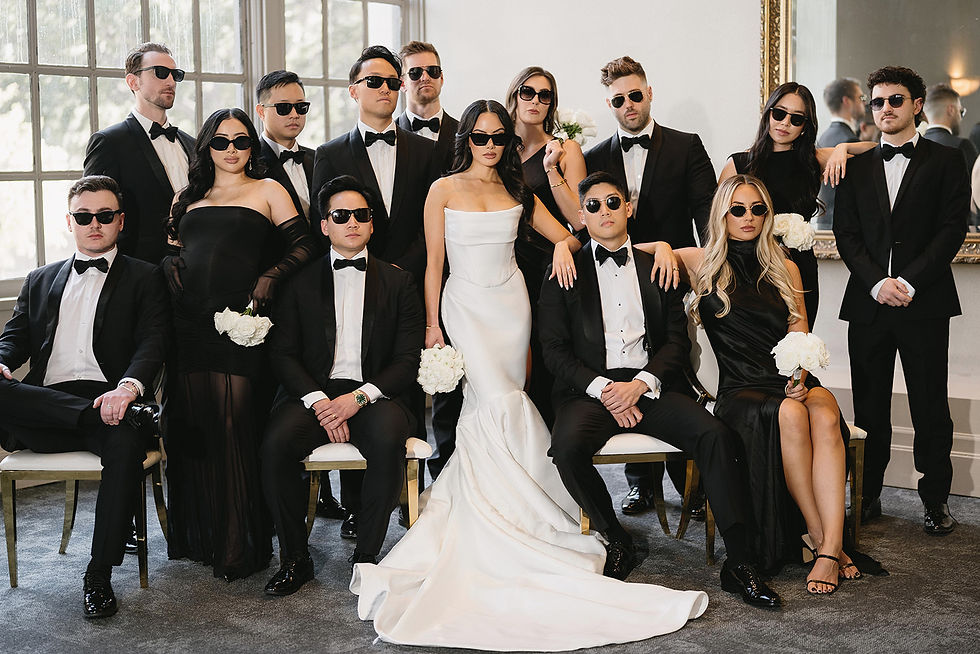 Wedding party posing indoors. The bride in white and guests in black tuxedos and dresses, all wearing sunglasses, hold white bouquets. Elegant mood.