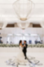 Bride and father of the bride dance surrounded by white flowers. Elegant hall with musicians in background and large chandeliers above, creating a romantic mood.