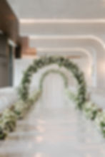 Elegant wedding aisle with white floral arches, surrounded by white chairs. Soft lighting creates a serene and romantic atmosphere.