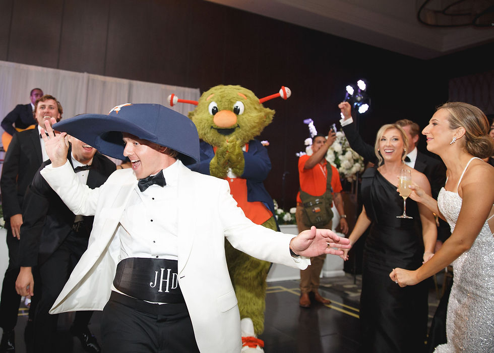 People dancing at a celebration, one in a white suit with a large blue hat. Houston Astros Orbit mascot claps in the background. Joyful atmosphere.
