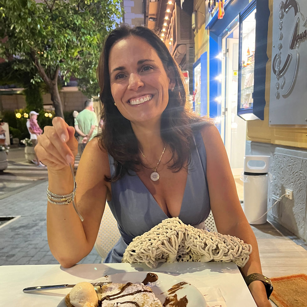 Woman in a blue dress smiles at a table with dessert outside a lit cafe. Trees and people in the background create a lively atmosphere.