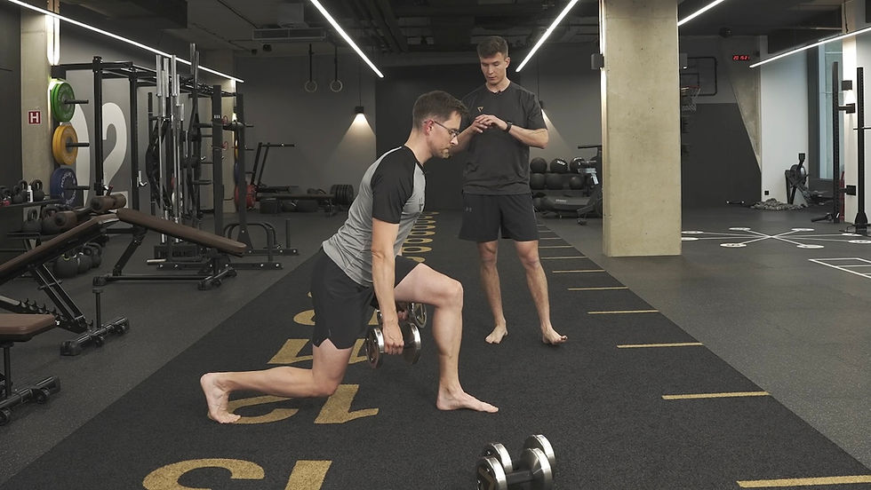 Muž provádí výpad s jednoručkami v moderní posilovně pod dohledem trenéra.