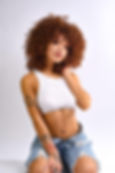 Honey Bxby poses in a studio portrait wearing a white crop top and denim shorts, showcasing her signature curls and tattoos.