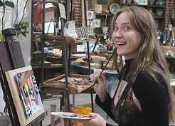 An artist is present in the gallery, shown working on a painting while people around her watch.