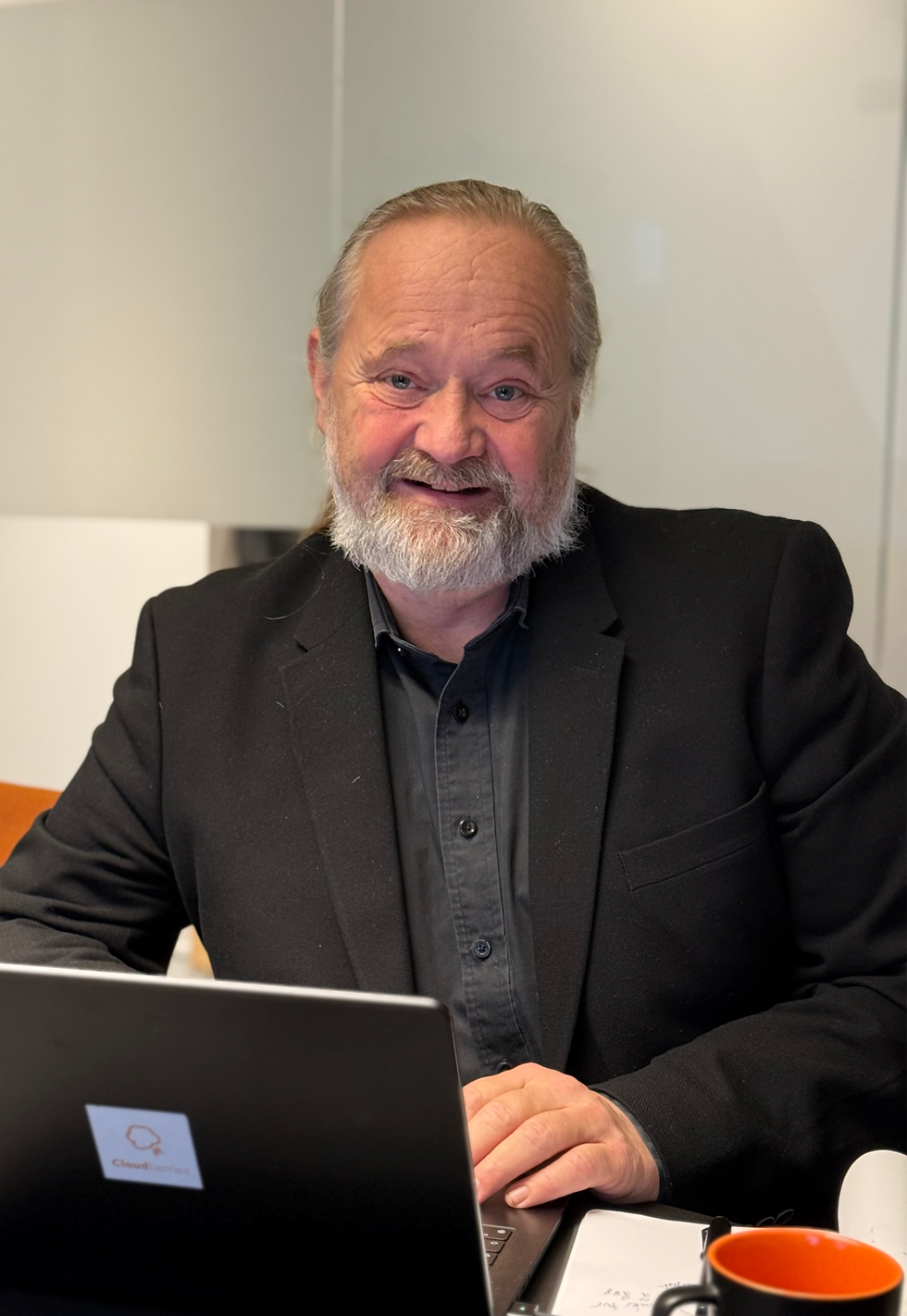 En mann med grått skjegg smiler til kameraet. Han har på seg svart dress og grå skjorte. Sitter foran en laptop og med en kaffekopp ved siden av. Kaffekoppen er svart med oransje farge inni.