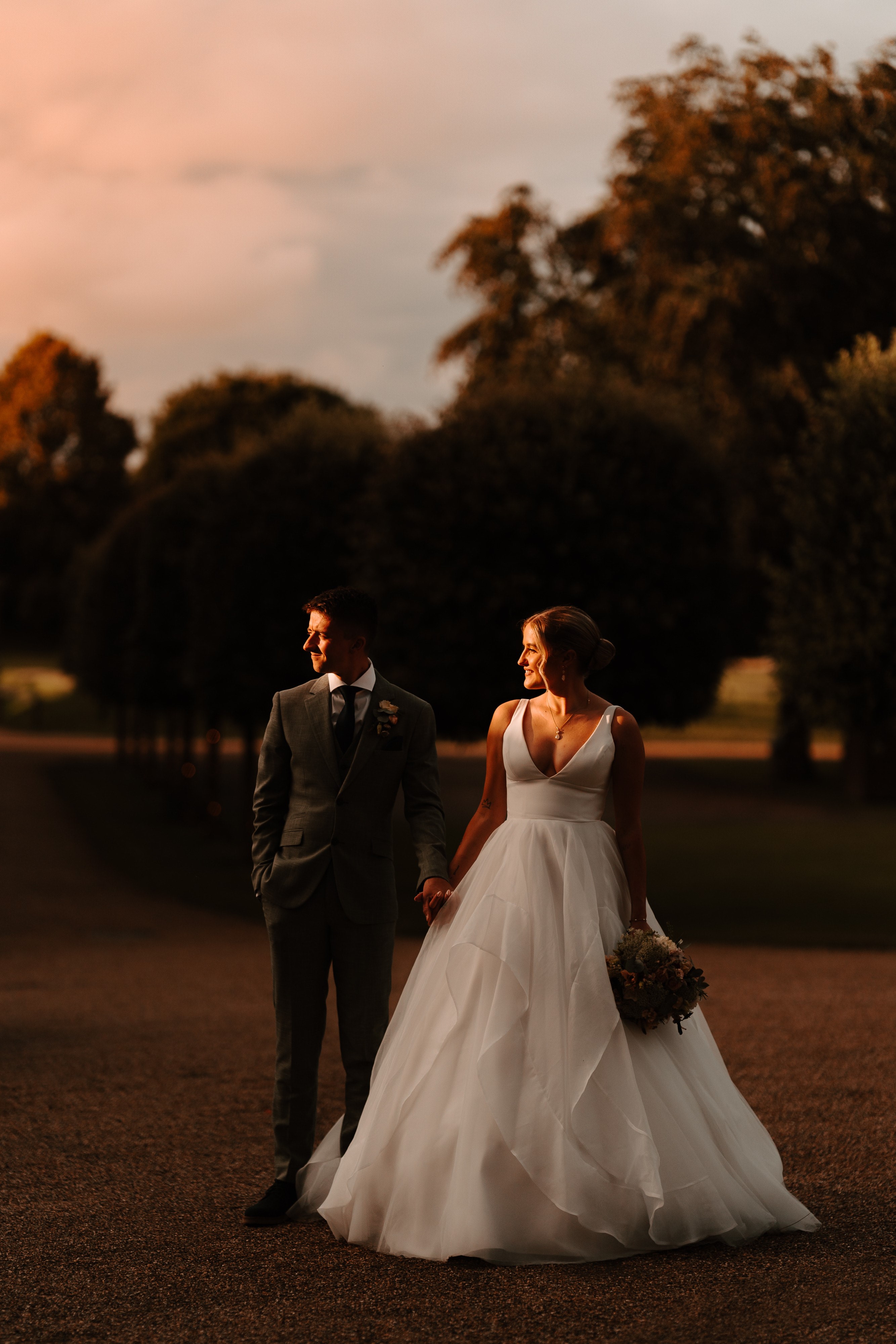 bride and groom holding hands whilst walking in field outside with evening sun casting shadows
