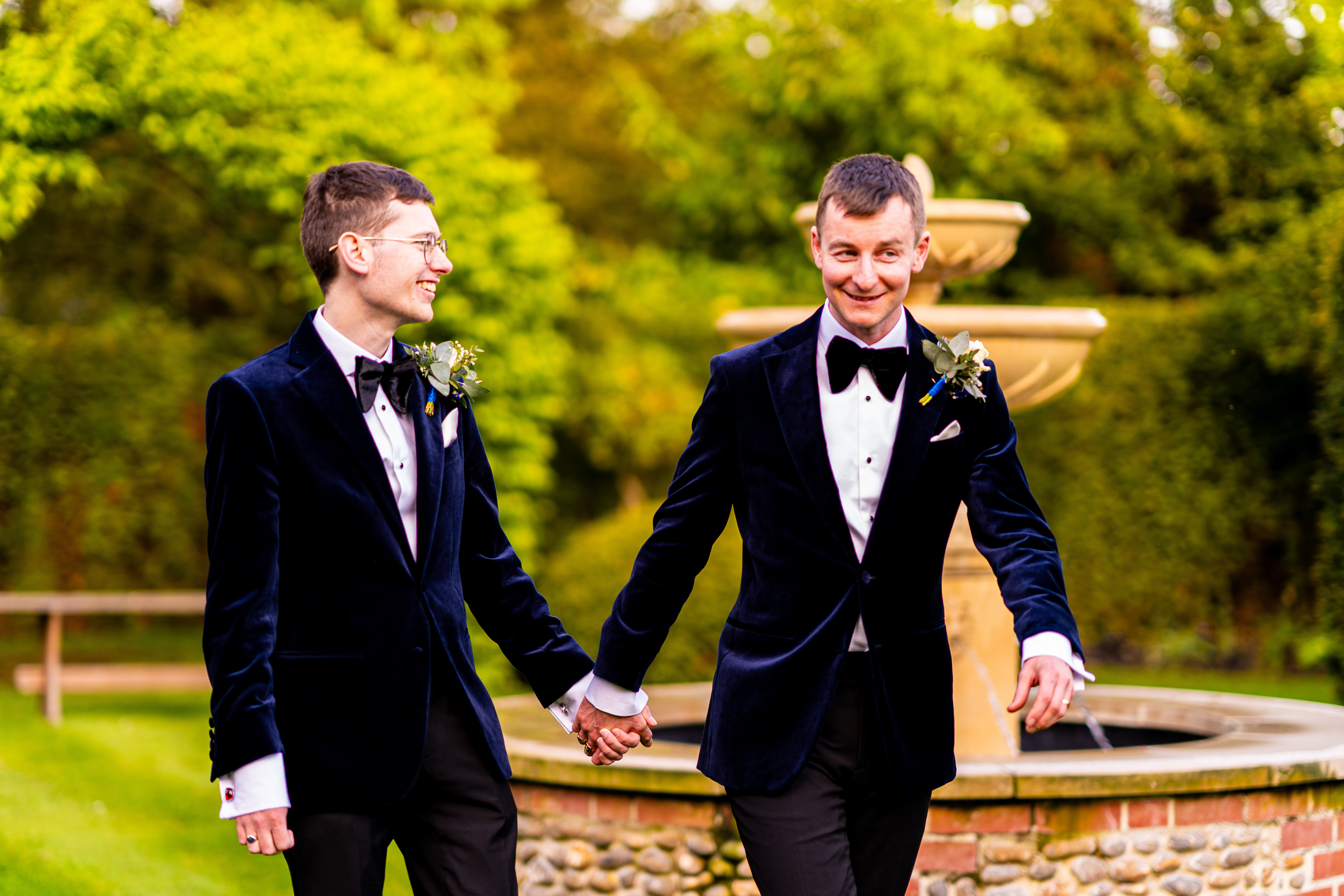 groom and groom holding hands and smiling in front of water fountain