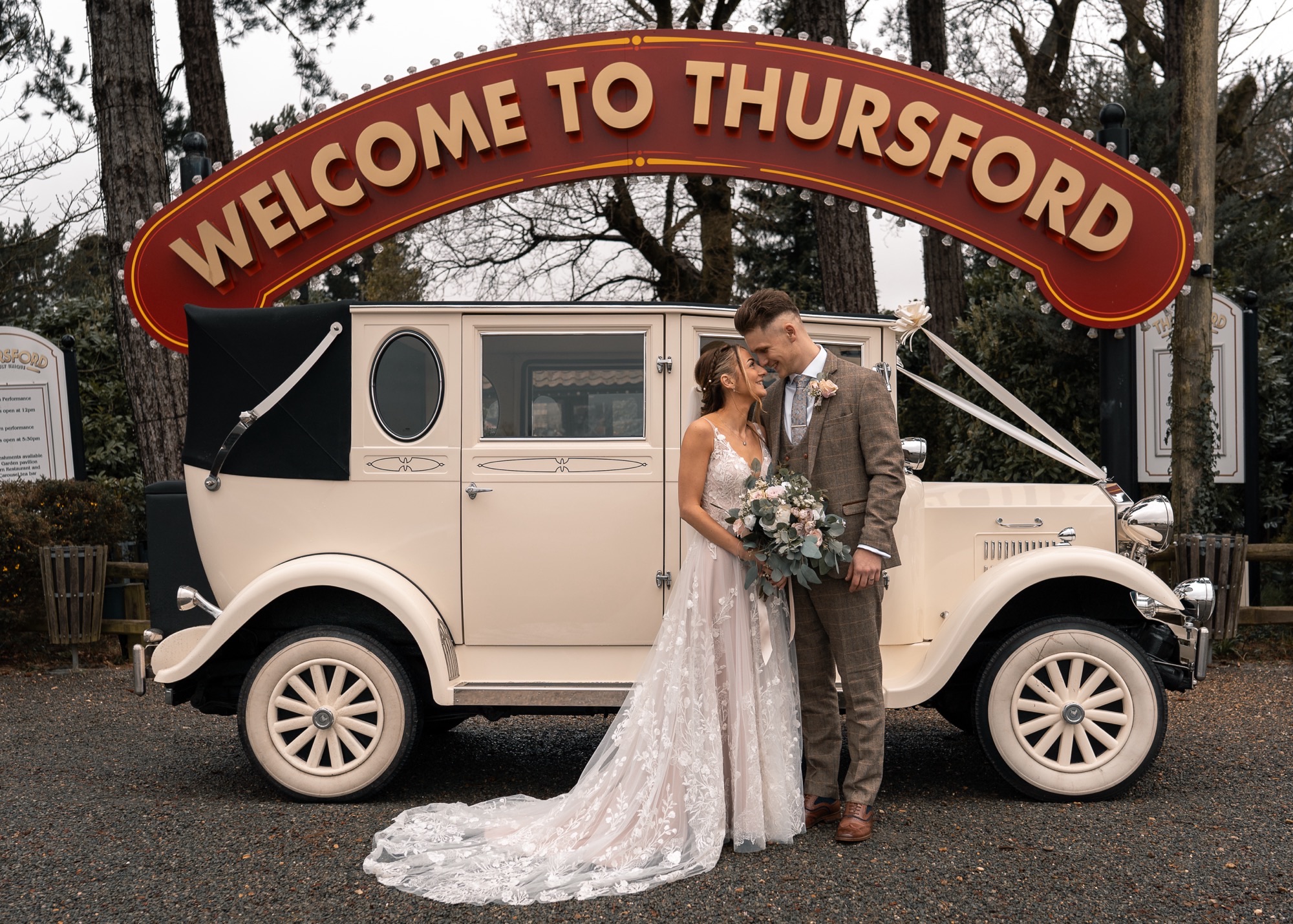 bride and groom hugging in front of old fashioned car with wedding bow with Thursford sign in the background