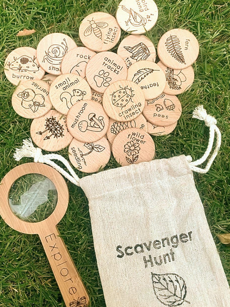 Small linen bag with the inscription "Scavenger Hunt", with wooden discs spilling out of it onto the lawn inscribed nature elements in words and images. A small wooden kids' magnifying glass lays beside it.