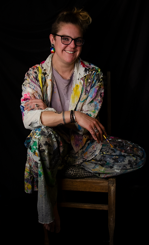 KatCoppock Headshot. Kat sits on a chair against a black background, wearing painted jeans and white shirt. She is wearing glasses and smiling, hair in a messy bun. She is wearing colour-wheel earrings and holding a paintbrush.