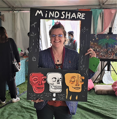 Kat stands smiling under an event marquee in a park, wearing a shimmery blue jacket and holding a frame around them that says 'Mindshare' and is illustrated with skulls