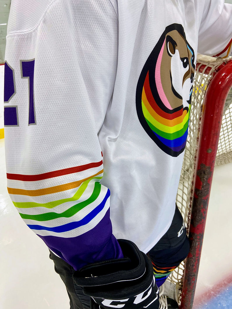 A close up of a hockey player standing by a net. The white jersey has rainbow stripes down the side.