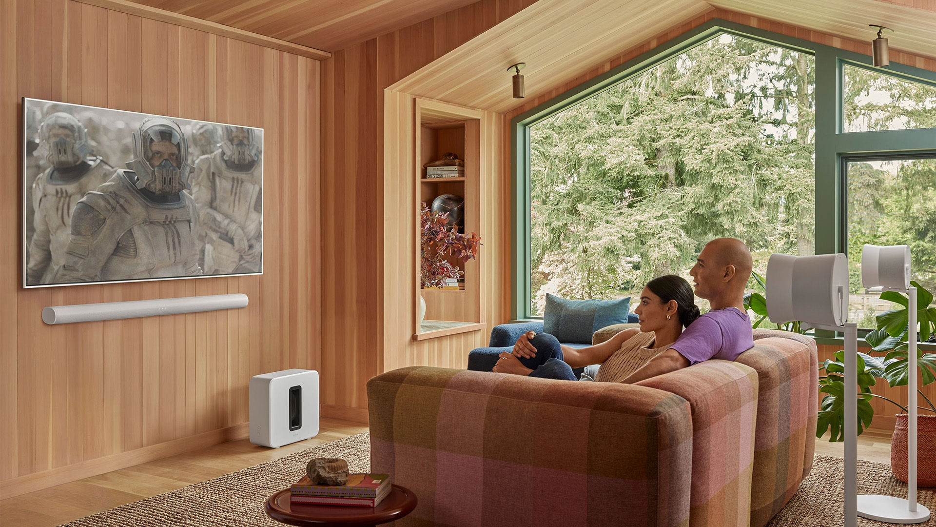 Couple watches TV, Sonos soundbar and speakers; wooden interior decor.