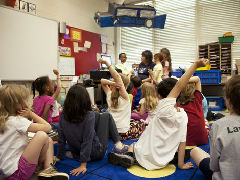An indoor classroom scene from a low angle showing children seated on the floor, some with hands raised, looking toward a teacher and student standing at the front during a lesson