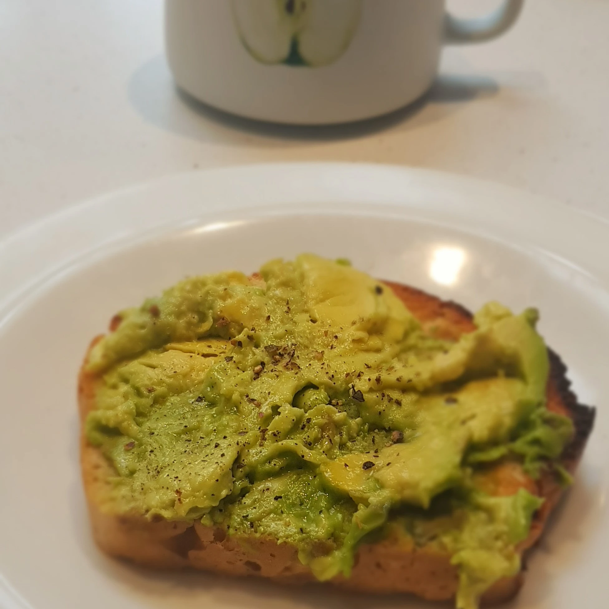 A slice of toast with smashed avocado on top, and a cup of coffee in the background
