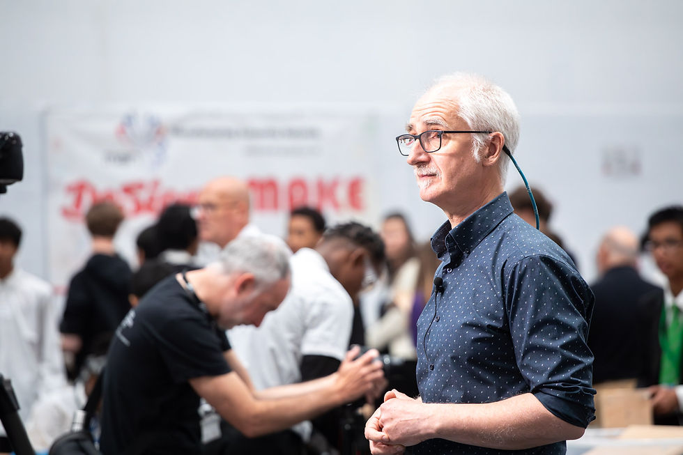 Elderly man in a blue shirt speaks with an engaged audience in the background. A banner with red text and blurred figures are visible.