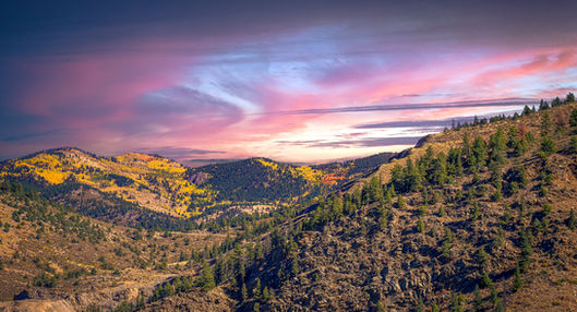 Dramatic mountain landscape at sunset featuring golden aspen trees on distant ridges, rugged rocky terrain, and a spectacular pink and purple sky creating a majestic twilight scene