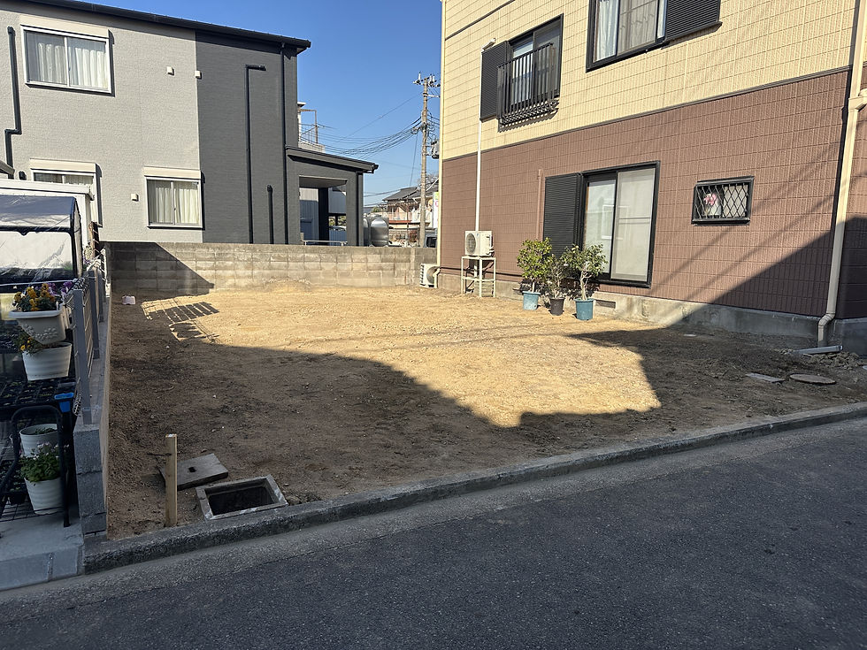 住宅街の空き地。砂地の更地が2階建て住宅に挟まれ、青空の下に植木鉢とエアコン室外機が並ぶ。