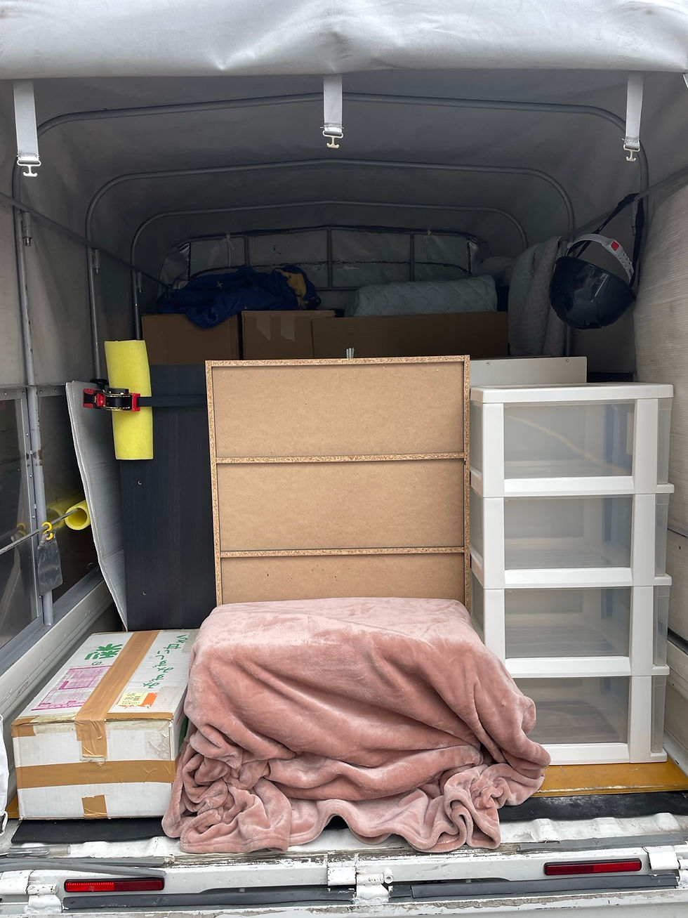 赤帽の幌付き軽トラックの荷台に、本棚、白い衣装ケース、段ボールなどの引っ越し荷物を積んだ様子。手前の荷物にはピンク色の毛布を掛けている。