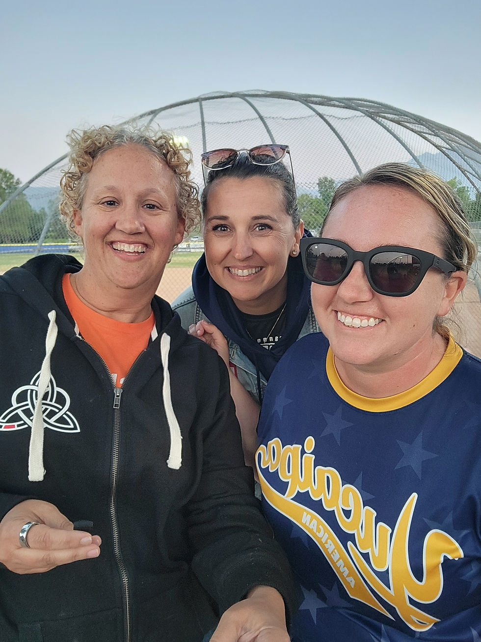3 smiling women, posed together, with a baseball field in the background
