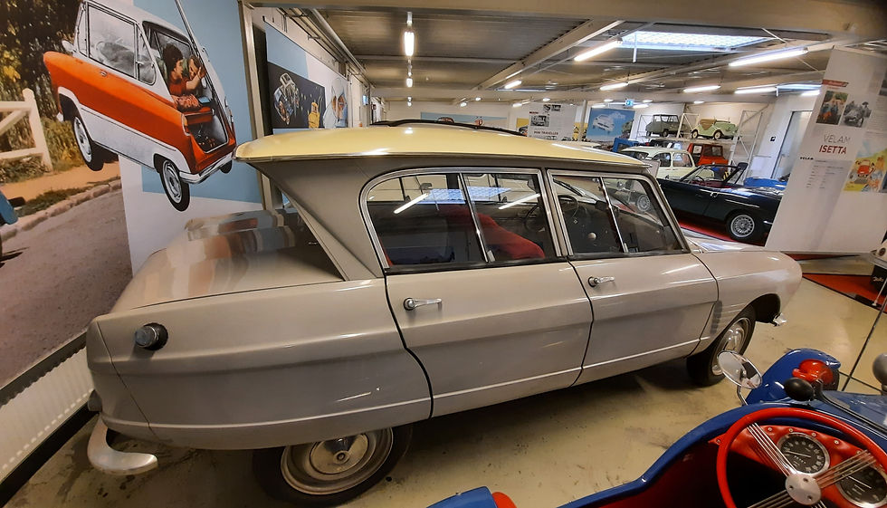 Silberfarbener Oldtimer mit cremefarbenem Dach und markantem Heck in einer Ausstellungshalle, im Hintergrund weitere historische Fahrzeuge.