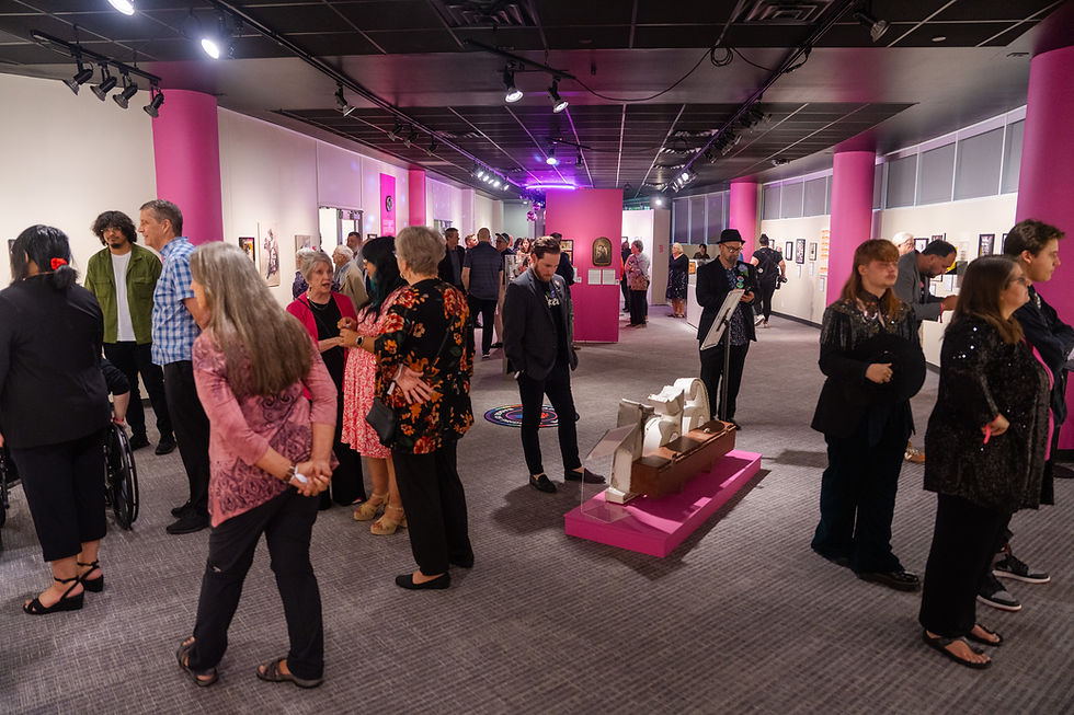 A wide view of a museum gallery filled with groups of visitors talking, reading exhibition labels, and examining artifacts. Bright pink columns and display walls extend through the space beneath a black ceiling with track lighting. A long artifact rests on a low pink platform at the center, while framed objects and labels line the surrounding walls. A wheelchair user appears on the left.