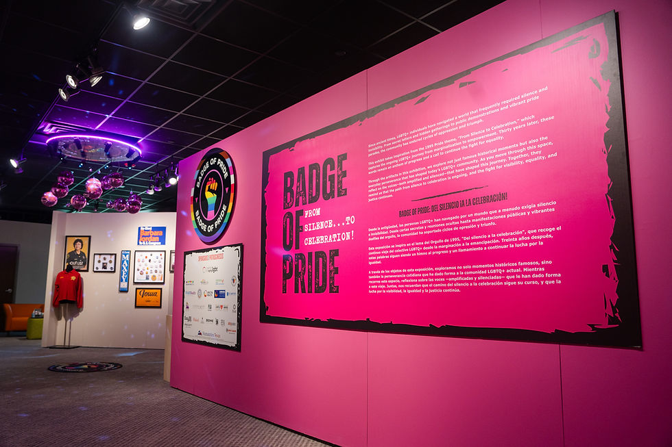 A wide view of the exhibition’s vibrant pink introductory wall. A large black title reads “Badge Of Pride,” with “From Silence…To Celebration!” in smaller white lettering. Several paragraphs of introductory text appear in English above a Spanish translation. An adjacent pink wall displays the circular Badge Of Pride logo and a panel recognizing exhibition sponsors. Pink mirrored disco balls hang from the black ceiling, and framed artifacts and a red shirt are visible in the gallery beyond.