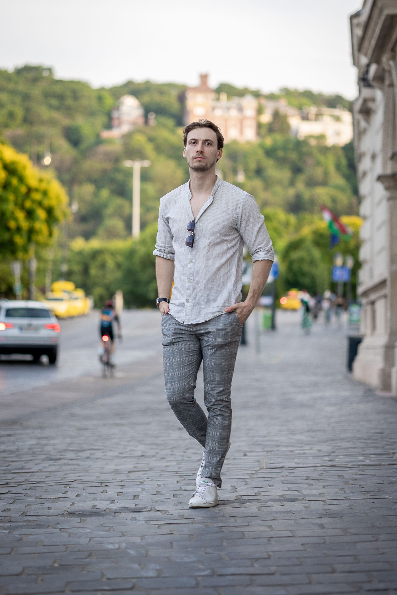 Férfi világos ingben, szürke kockás nadrágban és fehér cipőben áll egy macskaköves városi járdán.