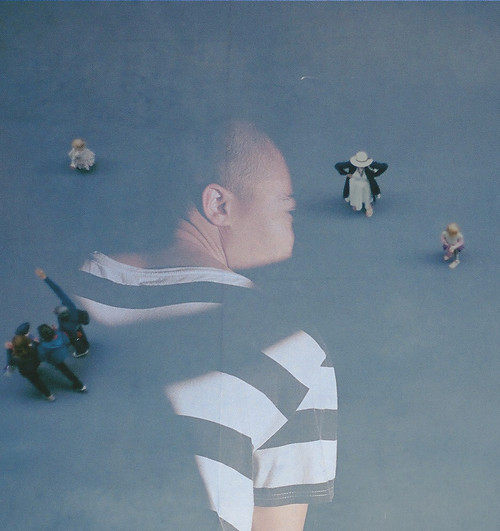 Two images are layered through one another. One image is a birds eye view of a large blue floored area. The centre top left of the image is a woman in a white dress, black coat and white hat with her arms on her sides, elbows jutting out. Far to the left is a girl toddler in a light coloured dress with blonde hair looking in the other direction. To the right, slightly below the woman is another young girl, also blond, in pink clothing, also looking to the left of the image. Centre bottom left are a group of three adults walking close together, one of them pointing, all looking in the same direction. The second image layered on top is of Oscar, He is larger than the others, taking up a third of the image, in the centre in between the other figures. He has his back to us but is turning his face so the side of his head is in view. He is in heavily contrasted light and shadow, only parts of him can be seen, his neck, eye, cheek, part of his black and white striped shirt and arm.