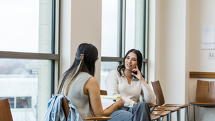 Two women sit and talk in a bright room with large windows. One holds notes, while the other listens, creating a thoughtful atmosphere.