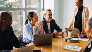 A diverse group of people smiling and discussing around a conference table with laptops and notes, in a bright room with large windows.