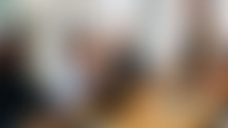 A diverse group of people smiling and discussing around a conference table with laptops and notes, in a bright room with large windows.