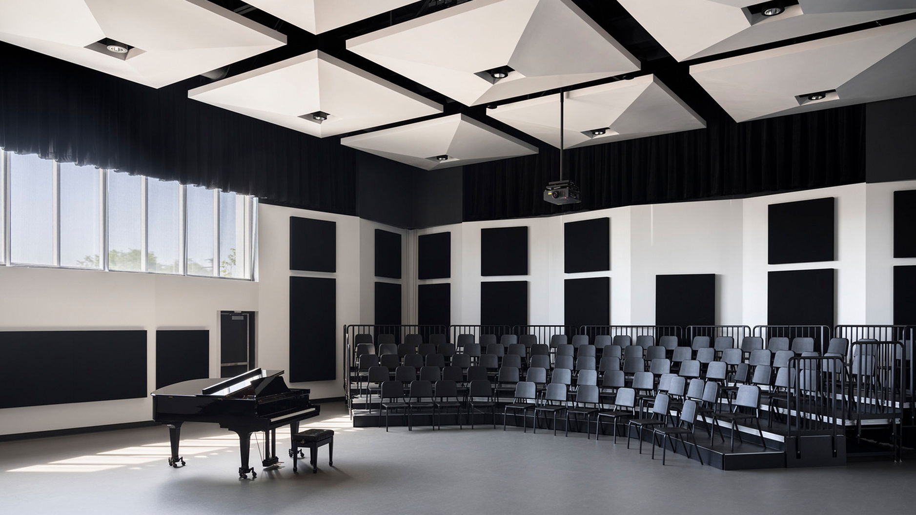 A photograph of the choir rehearsal room. The space has grey flooring, white walls with black sound dampening panels, a row of windows set above head-height, white geometric acoustic ceiling clouds, black choral risers with chairs, and a studio piano.