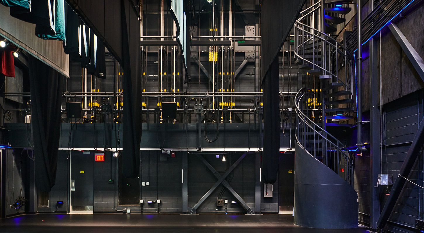 An interior view of the Mainstage Theatre at the Red Mountain Arts Campus. Looking from back stage towards the rigging. The venue is unoccupied and the house lights are on.