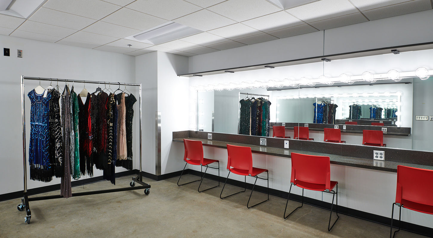 Photograph of one of the dressing rooms. The image shows a polished concrete floor, white walls, and a countertop with a long mirror, makeup lights, and red chairs. There is also a black z-rack with sequin costumes on it.