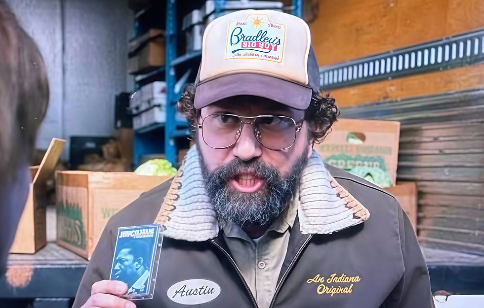 Man in glasses and hat holding a cassette tape, standing in a storage room with boxes. Jacket reads "Austin" and "An Indiana Original."