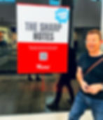 Man smiling next to a sign in a mall. The sign reads "THE SHARP NOTES," "COMING SOON," and "Westfield" with a QR code. Reflective floor.