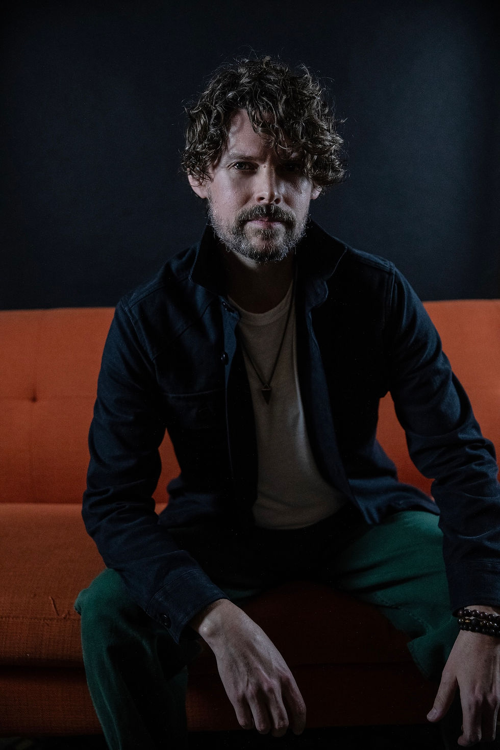 Man with curly hair sits on an orange couch, wearing a dark jacket and green pants, in a dimly lit room with a serious expression.