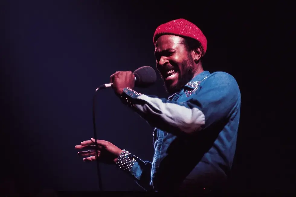 Singer in a red knit cap and blue shirt belts into a microphone on a dark stage, expressive and intense.