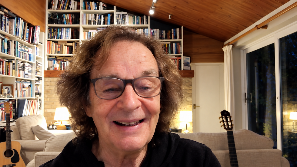 Smiling man with glasses in a cozy book-lined living room, with guitars, lamps, and a wood ceiling in the background