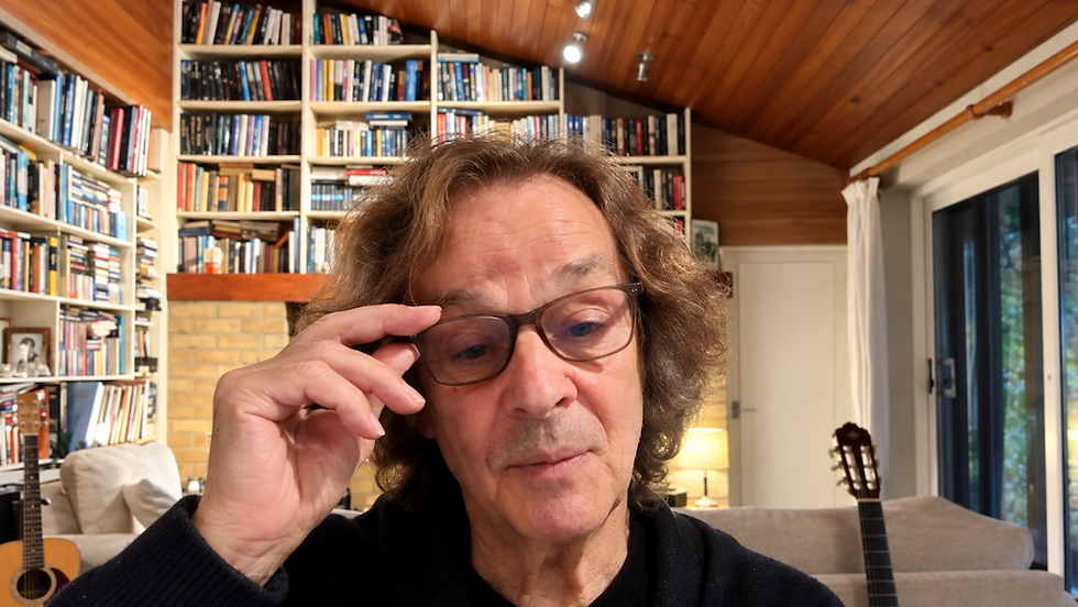 Middle-aged man adjusts his glasses in a cozy book-lined living room with warm wood ceilings, guitars, and a thoughtful expression.