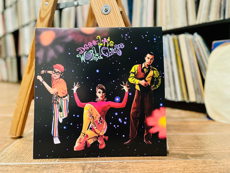 Album cover on a wooden easel in a record shop, showing three colorful performers and the text Deee-lite World Clique