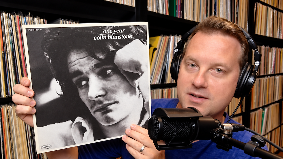 Man wearing headphones holds One Year by Colin Blunstone album cover in front of shelves of records, beside a microphone.