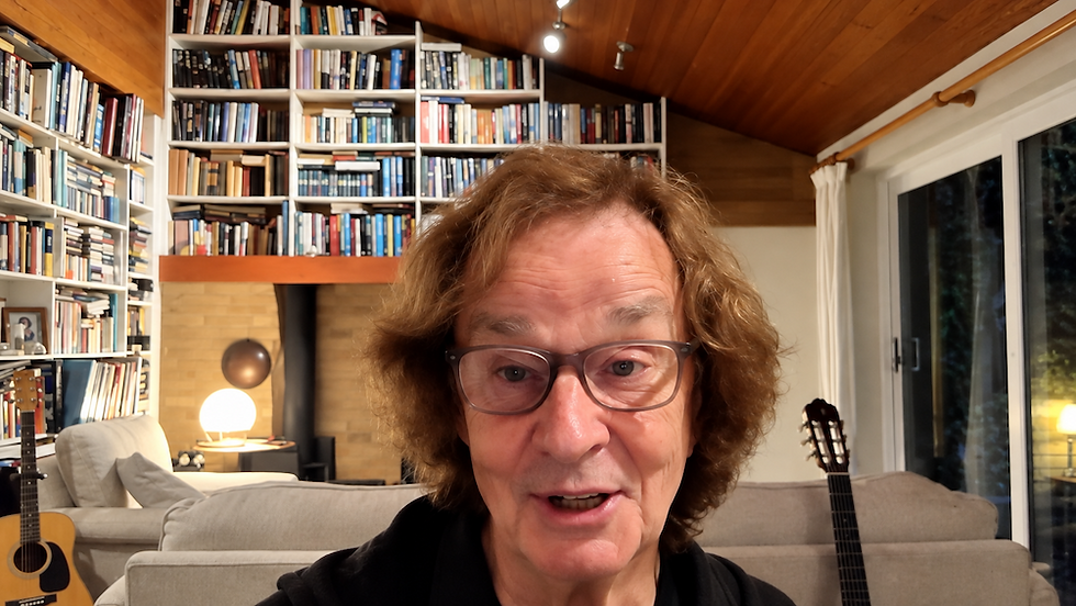 Man with glasses speaks in a cozy living room lined with bookshelves, guitars, and a lit lamp beneath a wood-paneled ceiling.