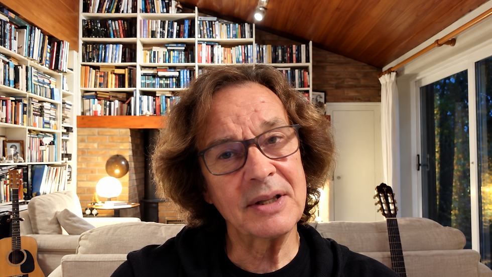 Colin Blunstone with glasses in a cozy book-lined living room, speaking to camera; guitars and warm lamp behind him.