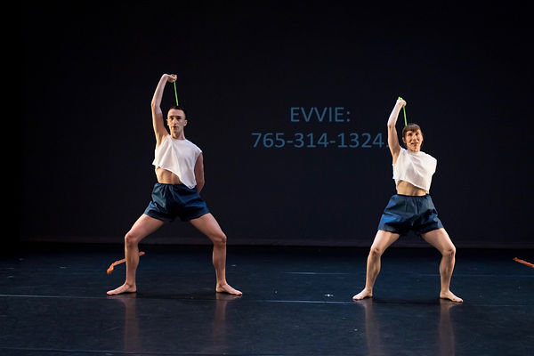 Elliot, a queer white person, and Evvie, a slim white woman, stand next to each other in a wide stance. They hold green thera-bands behind their heads, primed for an exercise. They stand on a stage where Evvie's phone number is posted behind them. They wear matching cropped, white shirts and papery blue pants.