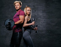 attractive-fitness-couple-sporty-male-holds-barbell-slim-blond-female-holds-dumbbells-grey