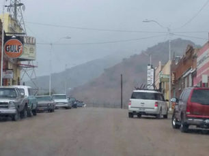 Moody, overcast view of Erie Street in Lowell, Bisbee during a monsoon, featuring a street lined with parked vehicles and vintage storefronts including a prominent Gulf gas station sign. The scene is enveloped in a hazy mist, adding a ghostly ambiance to this historic mining town.