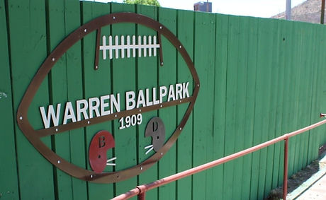Green wooden fence with wording that says "Warren Ballpark, 1909" in the shape of a football.
